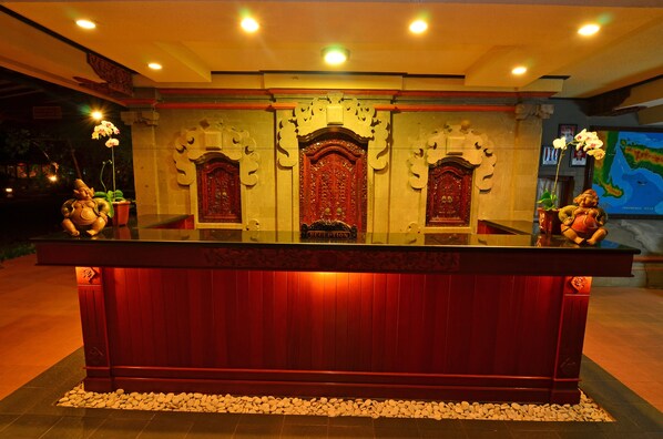A reception desk with ornate carvings and a red finish, flanked by decorative statues and a map on the wall. - Inna Sindhu Beach (Denpasar)
