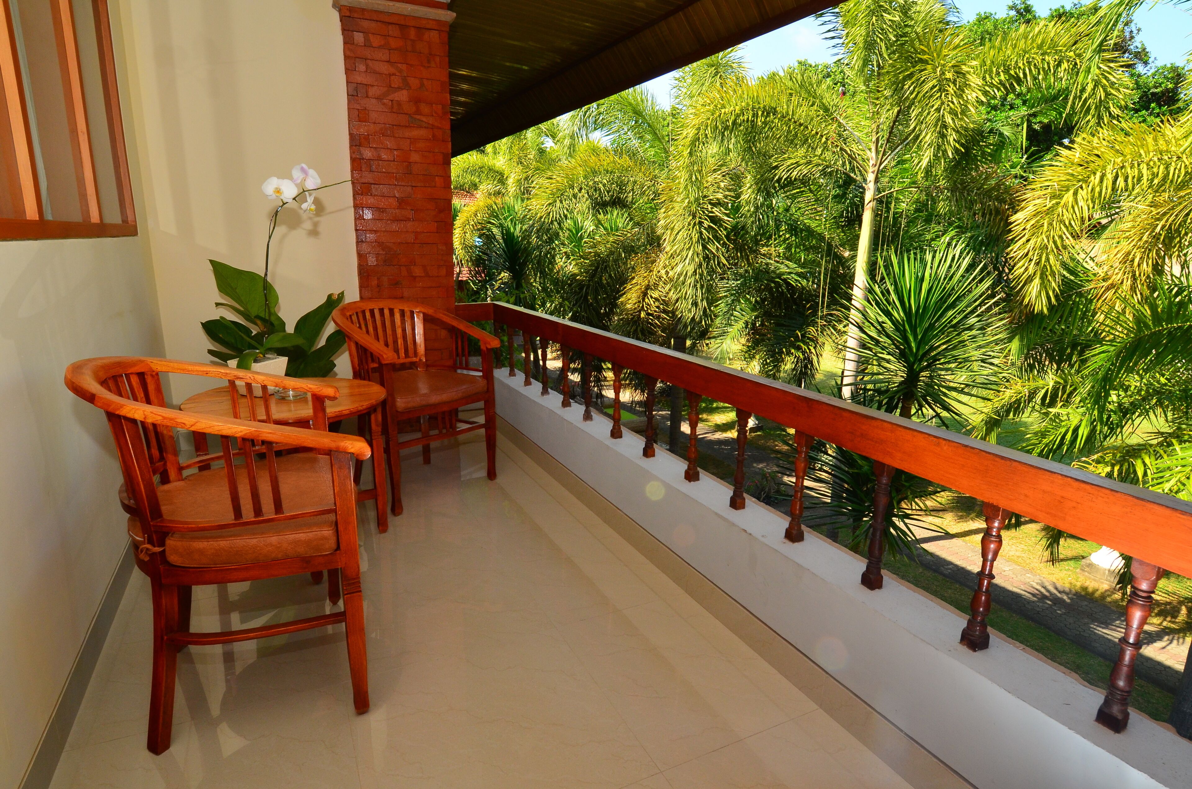 A balcony with wooden chairs and a table, overlooking a lush garden with palm trees.
