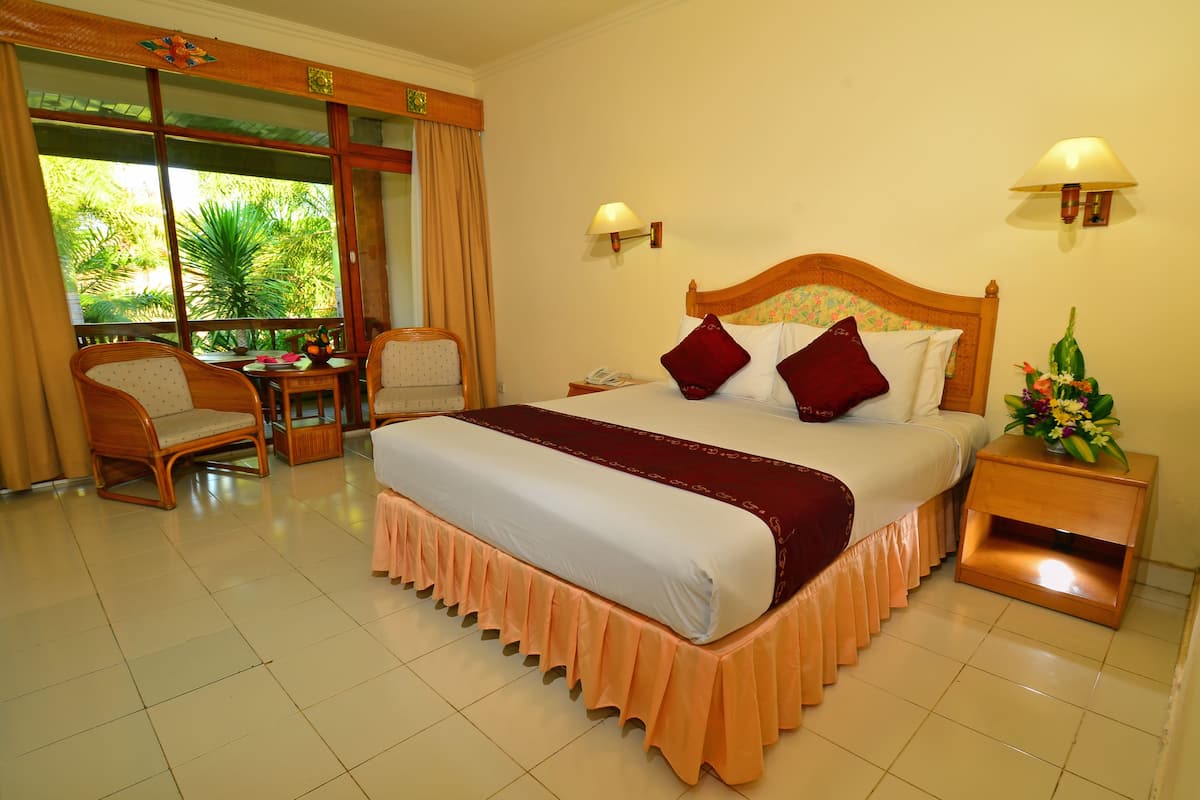 A hotel room with a large bed, two chairs, a small table, and a window with a view of greenery.