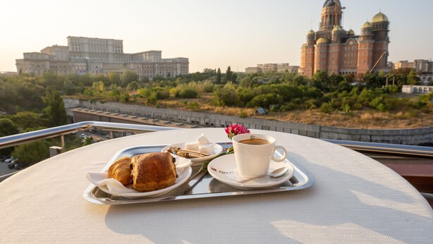 Una mesa con un croissant, una taza de café y una flor en un balcón con vistas a un paisaje urbano que incluye edificios históricos y una grúa de construcción.