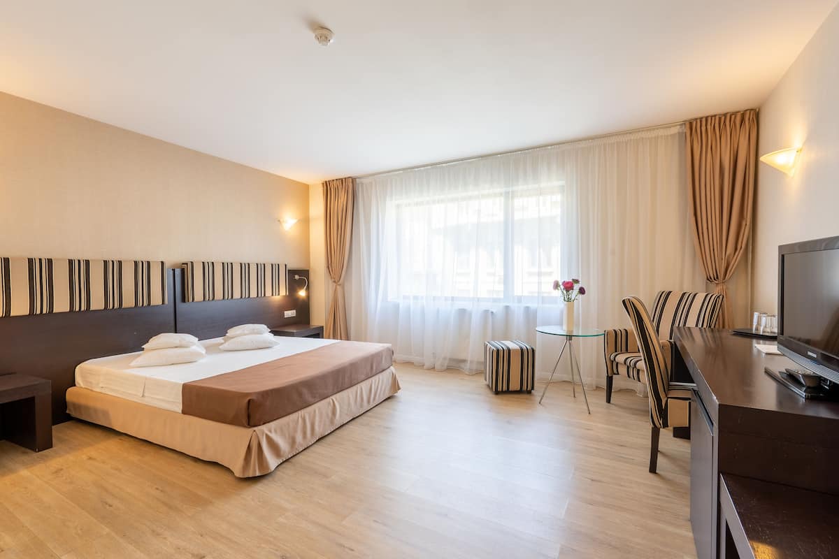 A hotel room with a bed, a desk with a TV, a chair, and a small table with a vase of flowers.