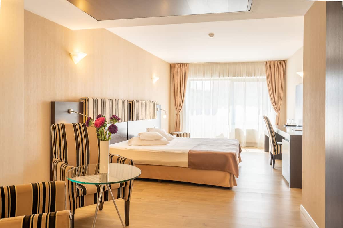 A hotel room with a bed, a glass table, striped armchairs, a desk, and a view of the outdoors.