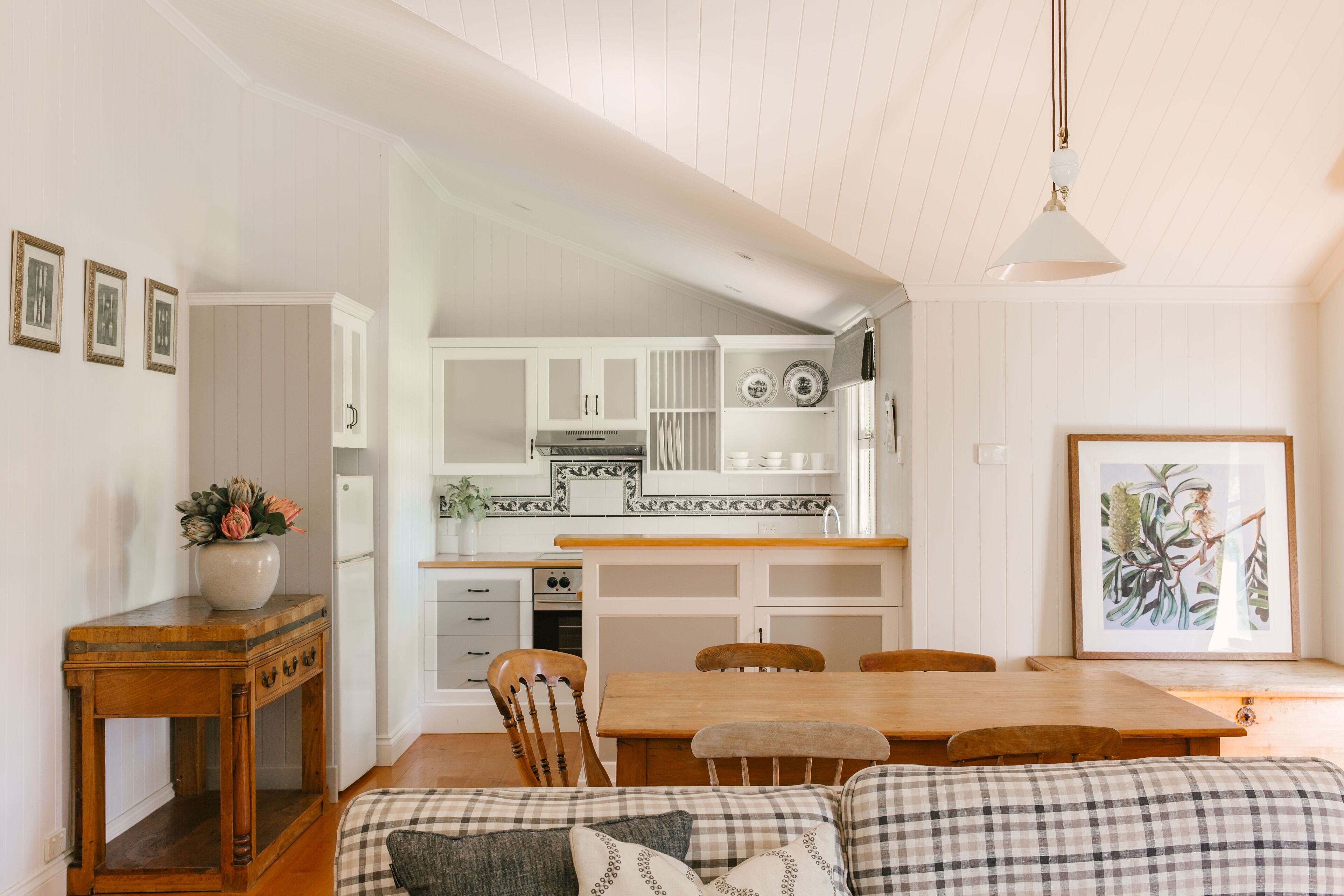 A cozy kitchen with wooden cabinets, a dining table, and chairs. There is a vase with flowers on a side table, and a framed picture on the wall.
