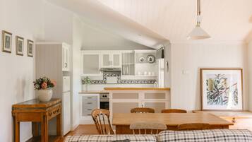 A cozy kitchen with wooden cabinets, a dining table, and chairs. There is a vase with flowers on a side table, and a framed picture on the wall.