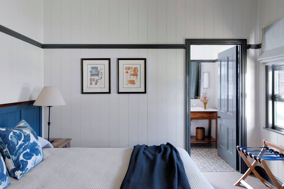 A hotel room with a bed, a nightstand, a lamp, a framed picture, a bathroom with a sink and mirror, and a window with blinds.