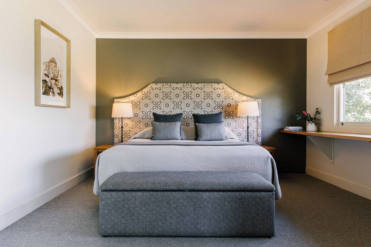 A neatly arranged bedroom with a patterned headboard, two bedside lamps, a bedside table with a book, and a window with a view.