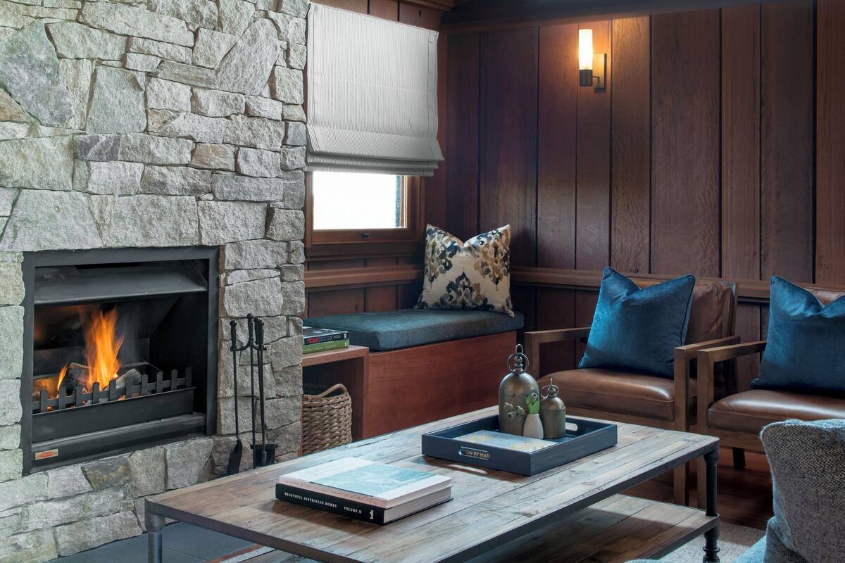 A cozy living room with a stone fireplace, wooden furniture, and a large window.