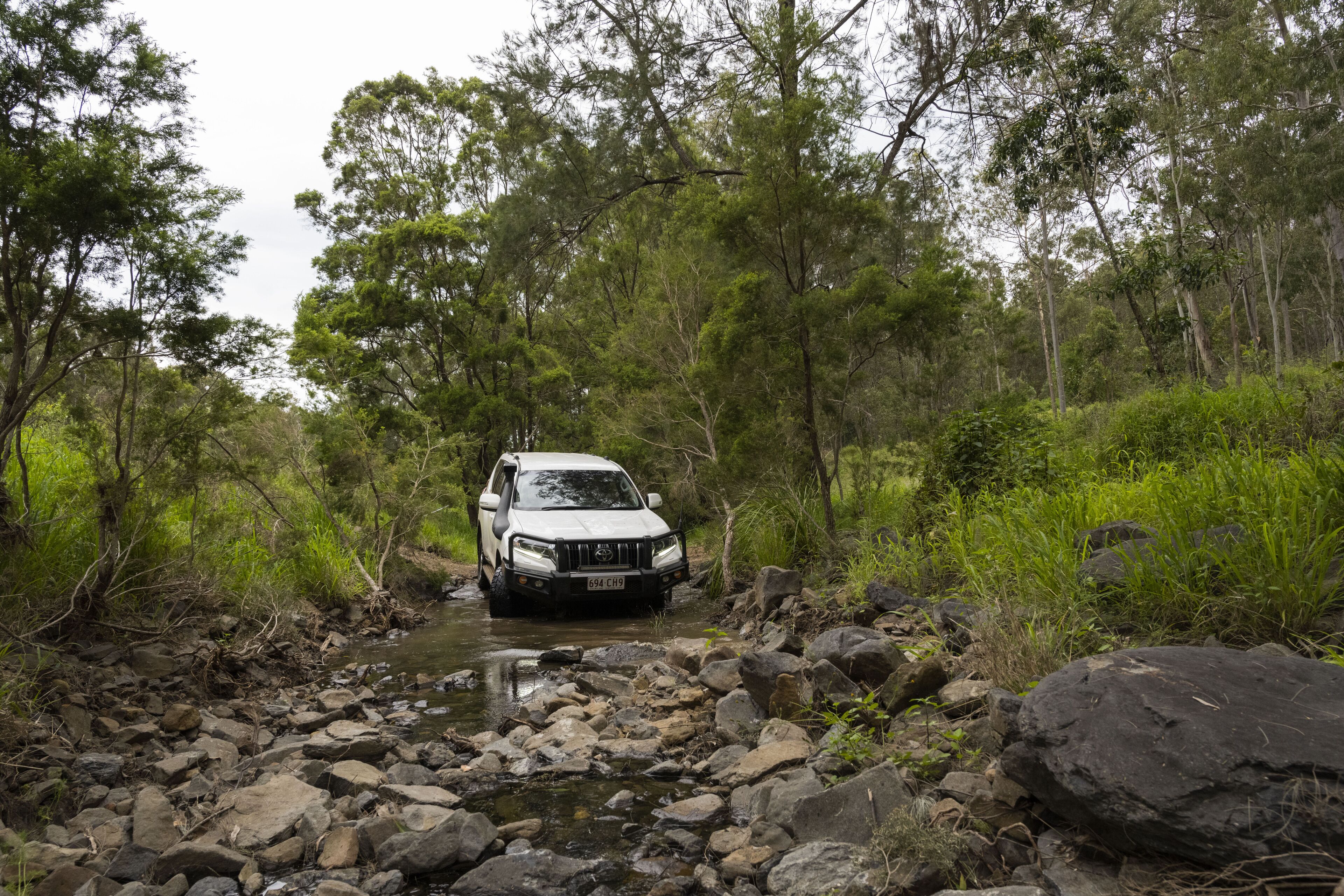 A vehicle navigating a rocky stream in a forested area.