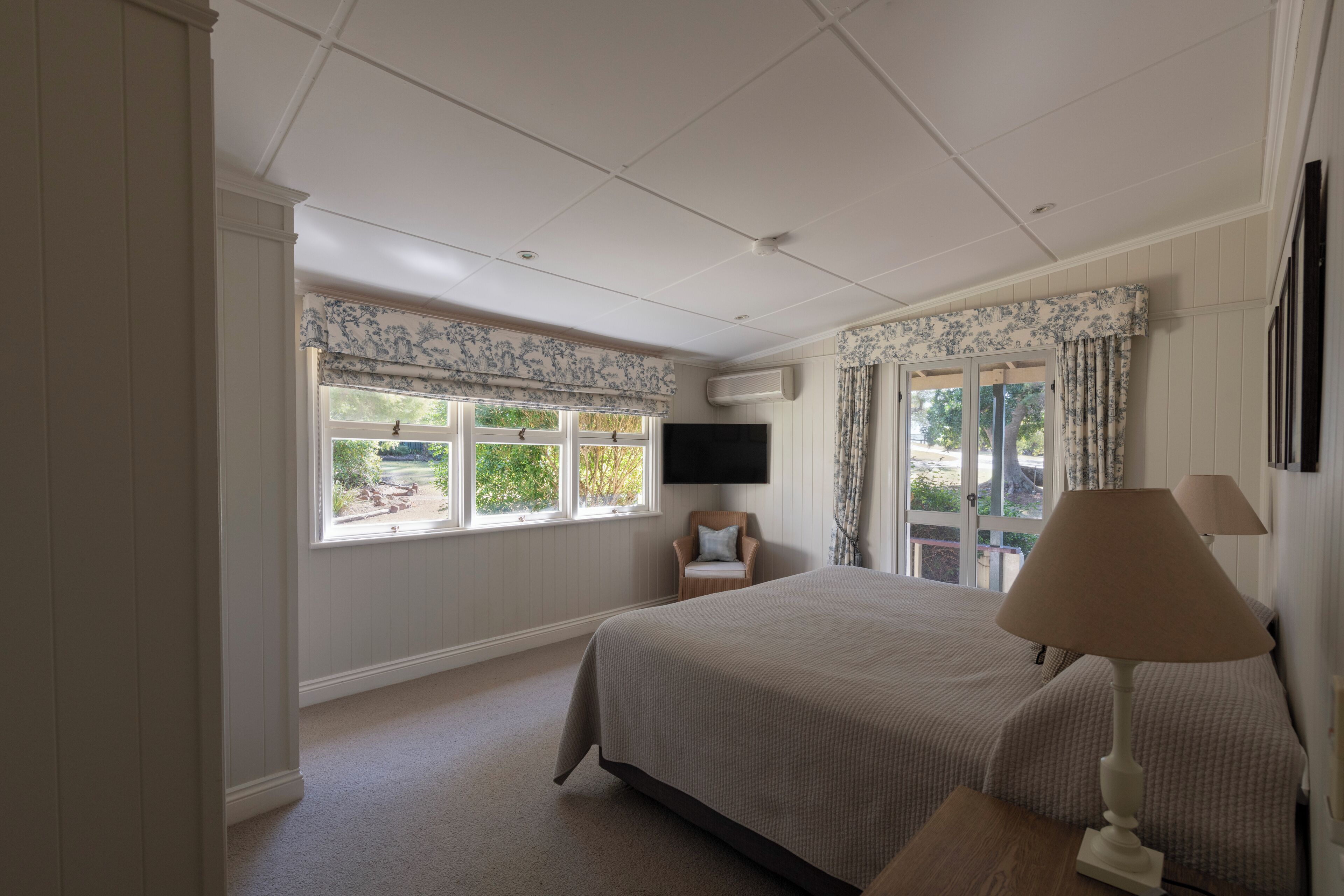 Un dormitorio con cama, televisor montado en la pared, ventana con vistas a la vegetación y puerta que da al exterior.