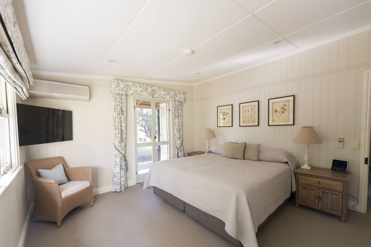 A bedroom with a bed, a chair, a nightstand, a TV, and a window with curtains.