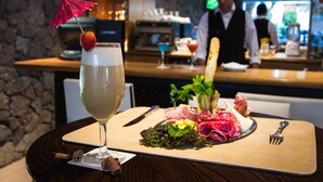 A cocktail with a strawberry garnish and a pink umbrella, a plate of assorted meats and cheese, and a fork on a wooden board.