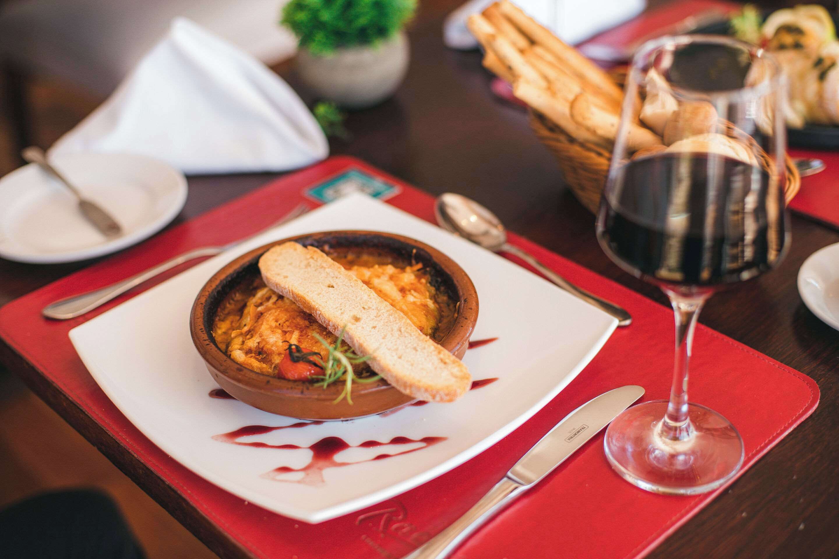 A dining table with a bowl of food, a glass of red wine, and bread on a red placemat.