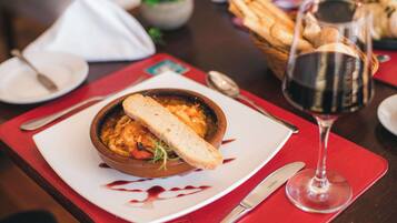 A dining table with a bowl of food, a glass of red wine, and bread on a red placemat.