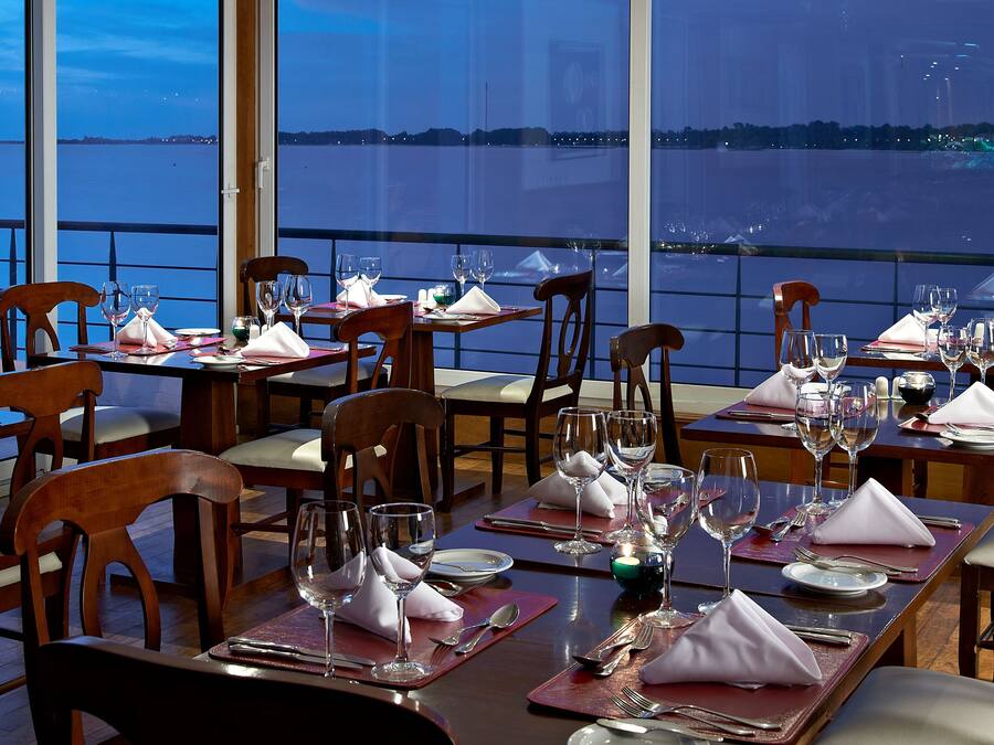 A dining area with tables set for multiple guests, each with wine glasses, plates, and cutlery. The setting features wooden chairs and a view of a body of water through large windows.