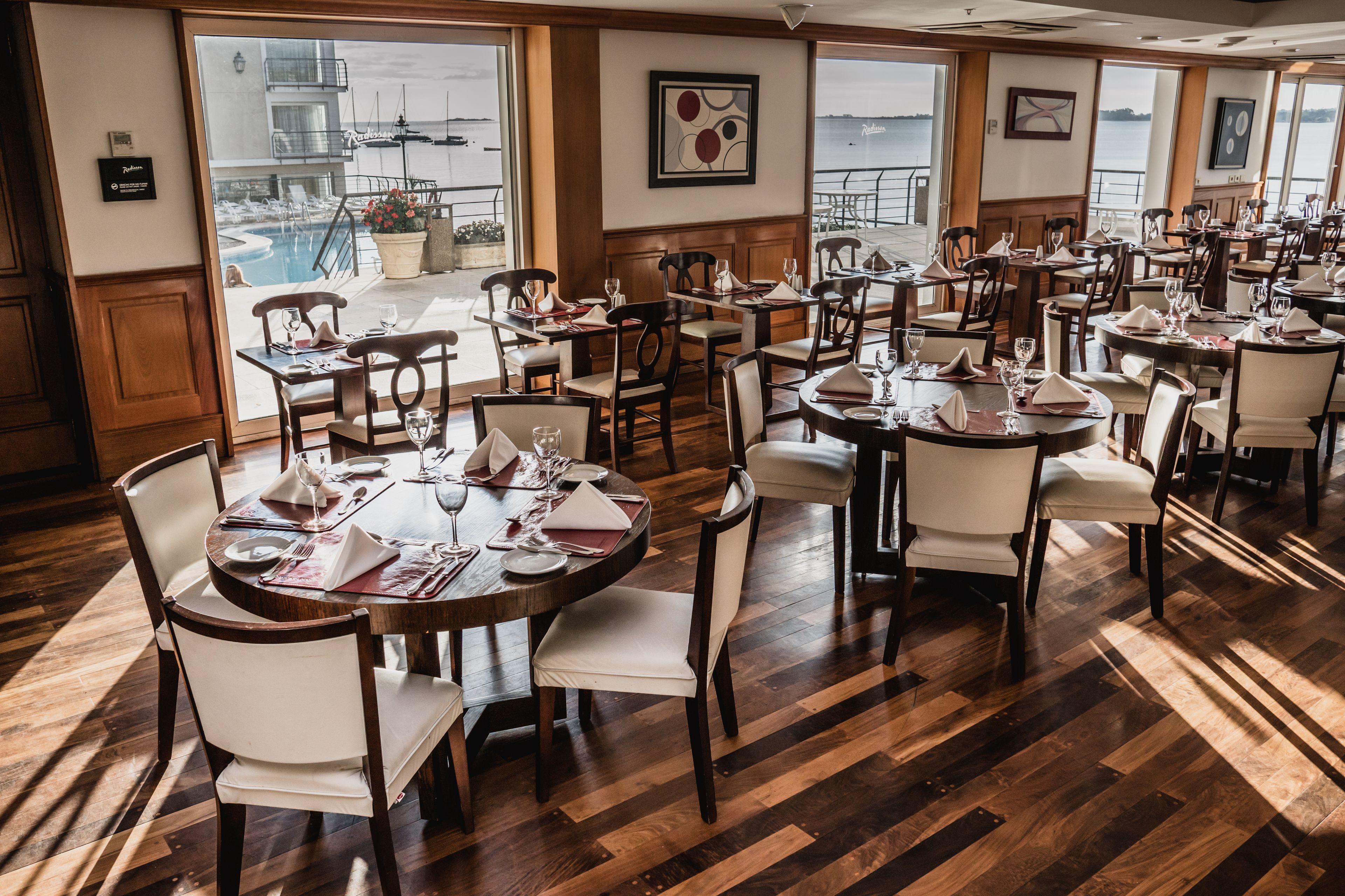 Un restaurant doté de nombreuses tables et chaises, de grandes fenêtres offrant une vue sur un quai et des bateaux, et d’un plancher en bois.