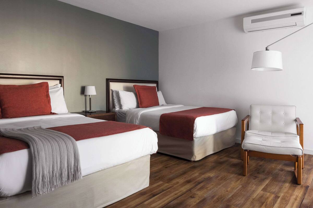 Hotellihuone, jossa on kaksi sänkyä, tuoli, lamppu ja ilmastointilaite.