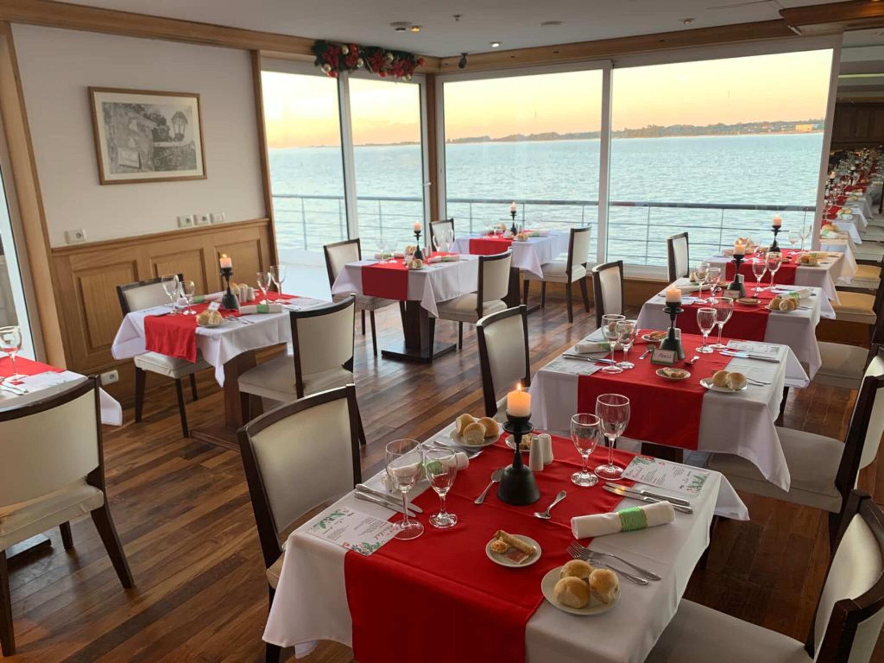 A dining area with tables set for a meal, large windows with a view of the sea, and a framed picture on the wall.