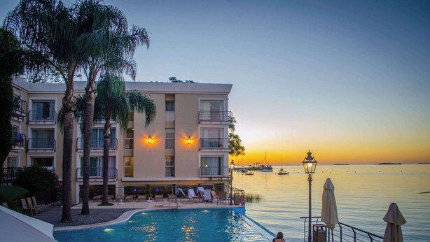 A hotel with a pool, lounge chairs, and a view of the sea at sunset.