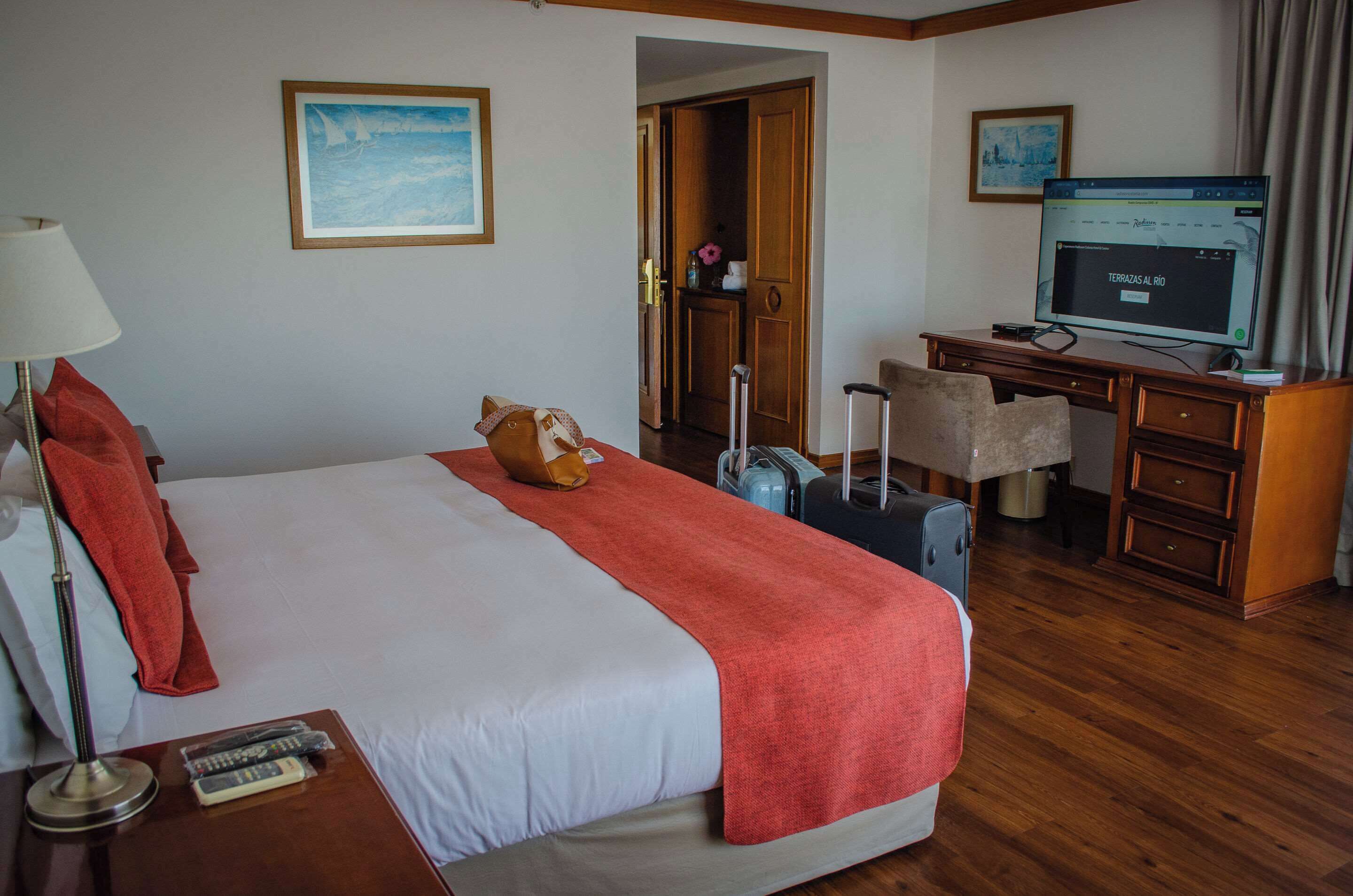 A hotel room with a bed, a desk, a chair, a television, and a suitcase.