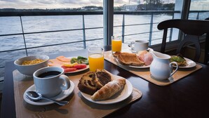 A breakfast table with a variety of food and drinks, a view of the water and sky through large windows.