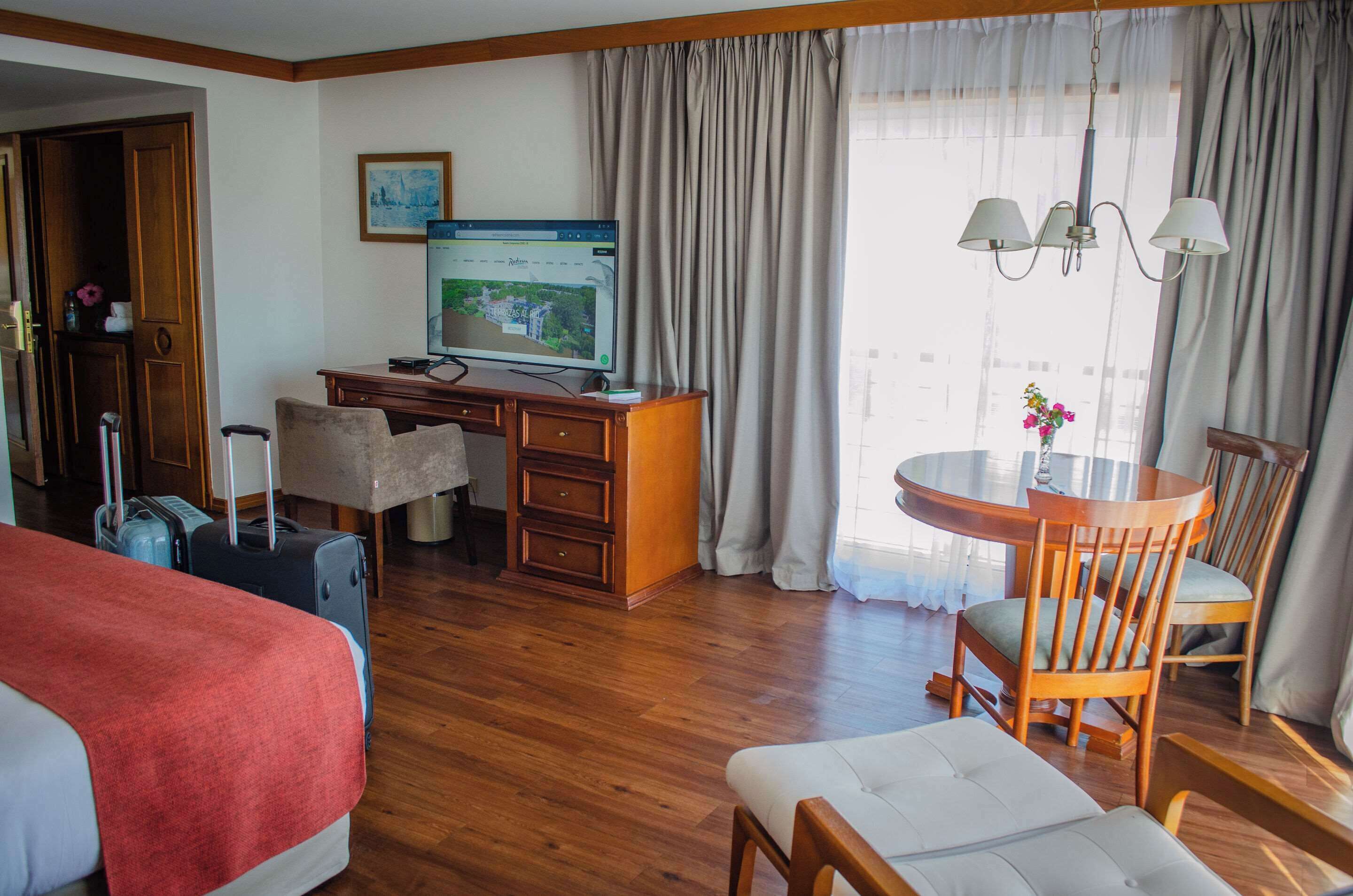Een hotelkamer met een houten bureau, een computer, een bed, een eettafel met stoelen en een raam met gordijnen.