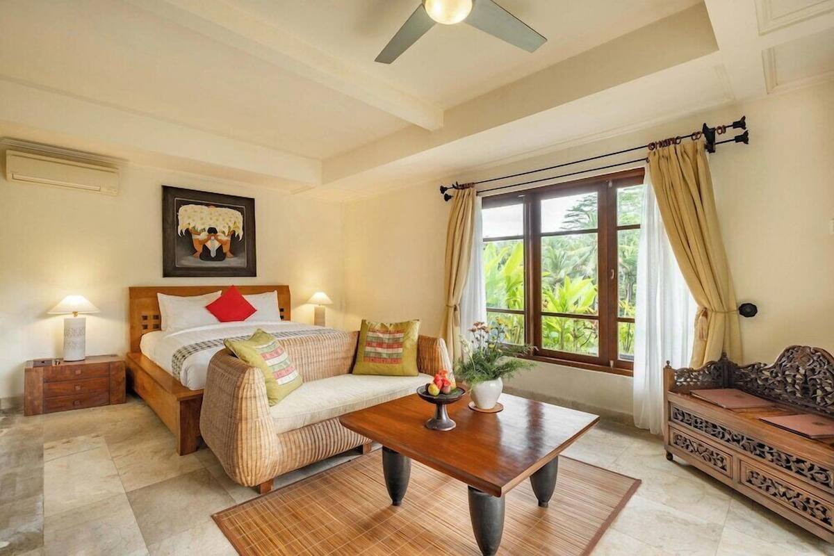 A bedroom with a bed, a sofa, a coffee table, a nightstand, a ceiling fan, and a window with curtains.