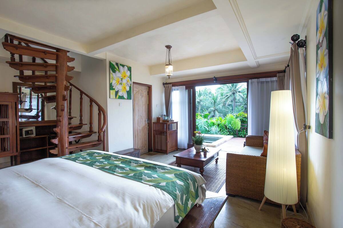 A bedroom with a bed, wooden staircase, a small table, a lamp, and a view of a garden through a sliding door.