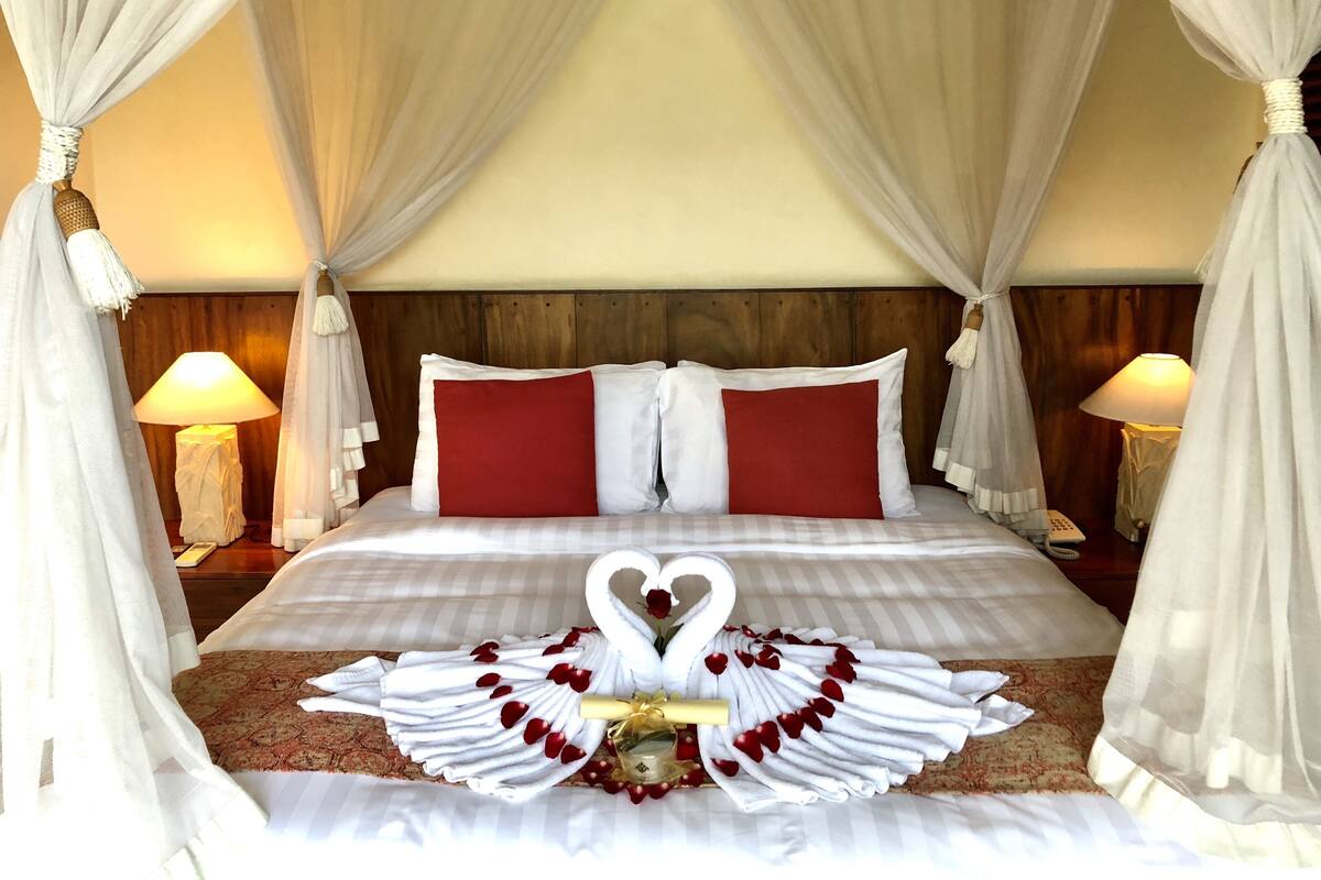 A bed with a canopy, two bedside lamps, and a swan decoration made of towels and rose petals.