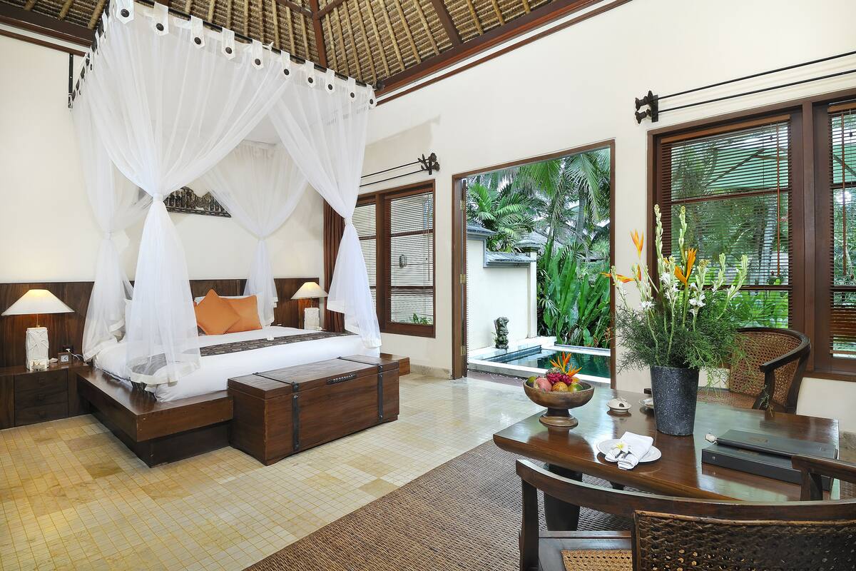 A bedroom with a four-poster bed, a wooden nightstand, a bedside table, and a view of a swimming pool and lush greenery.