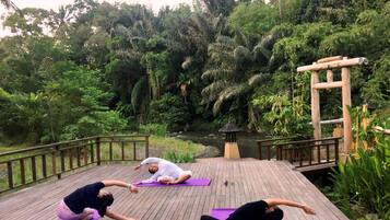 Una terraza de madera con tres personas practicando yoga sobre esterillas moradas, rodeado de exuberante vegetación y un pequeño estanque.
