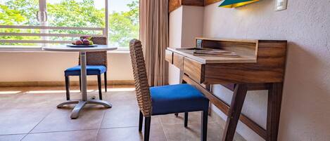 Una habitación con un escritorio de madera, una silla, una pequeña mesa con fruta y vistas a la vegetación a través de una ventana.