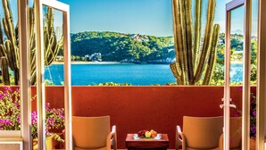 Un balcón con dos sillas, una mesa con fruta y vistas a un paisaje costero con cactus y edificios.