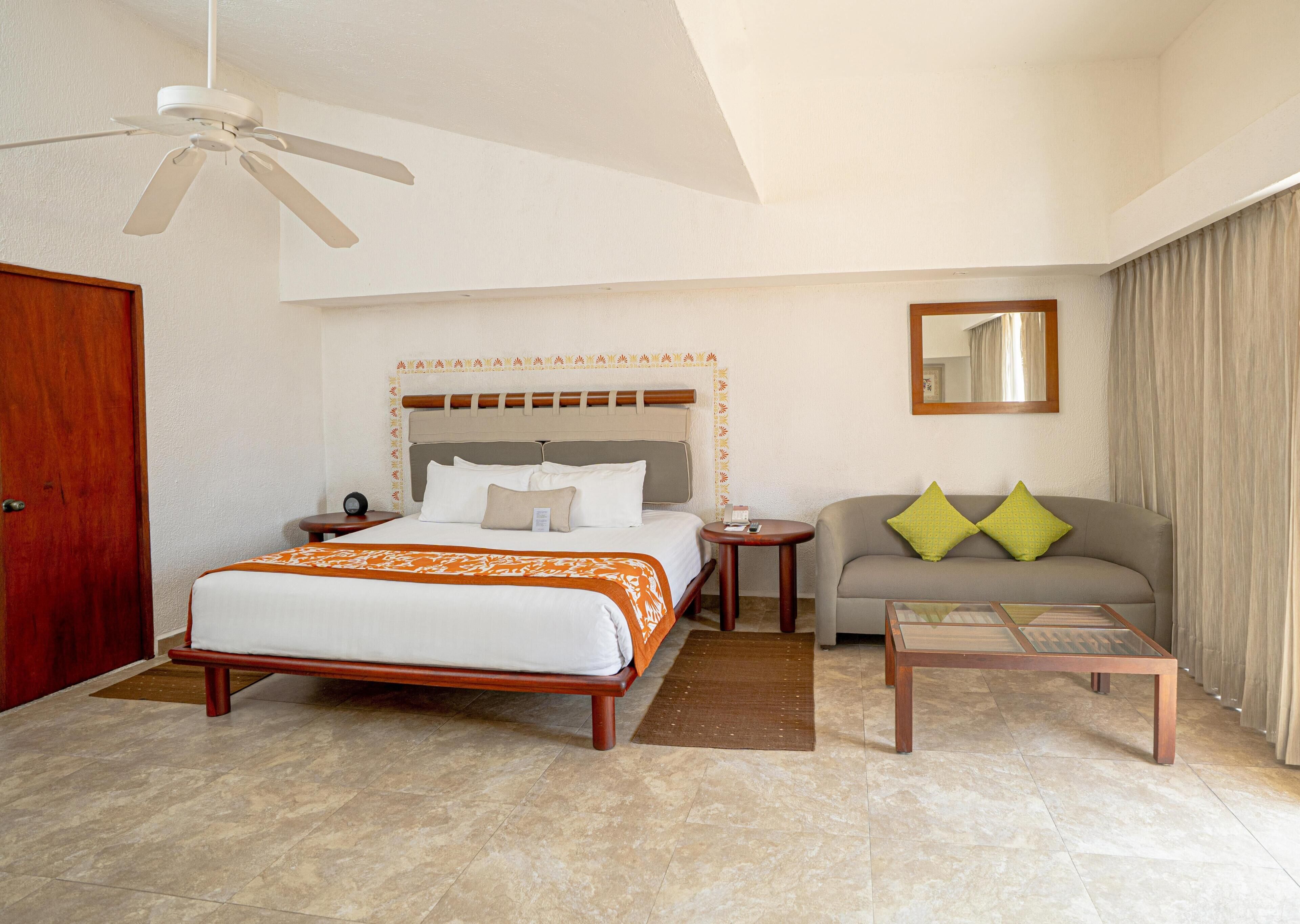 A hotel room with a bed, a sofa, a coffee table, a ceiling fan, a mirror, and a door.