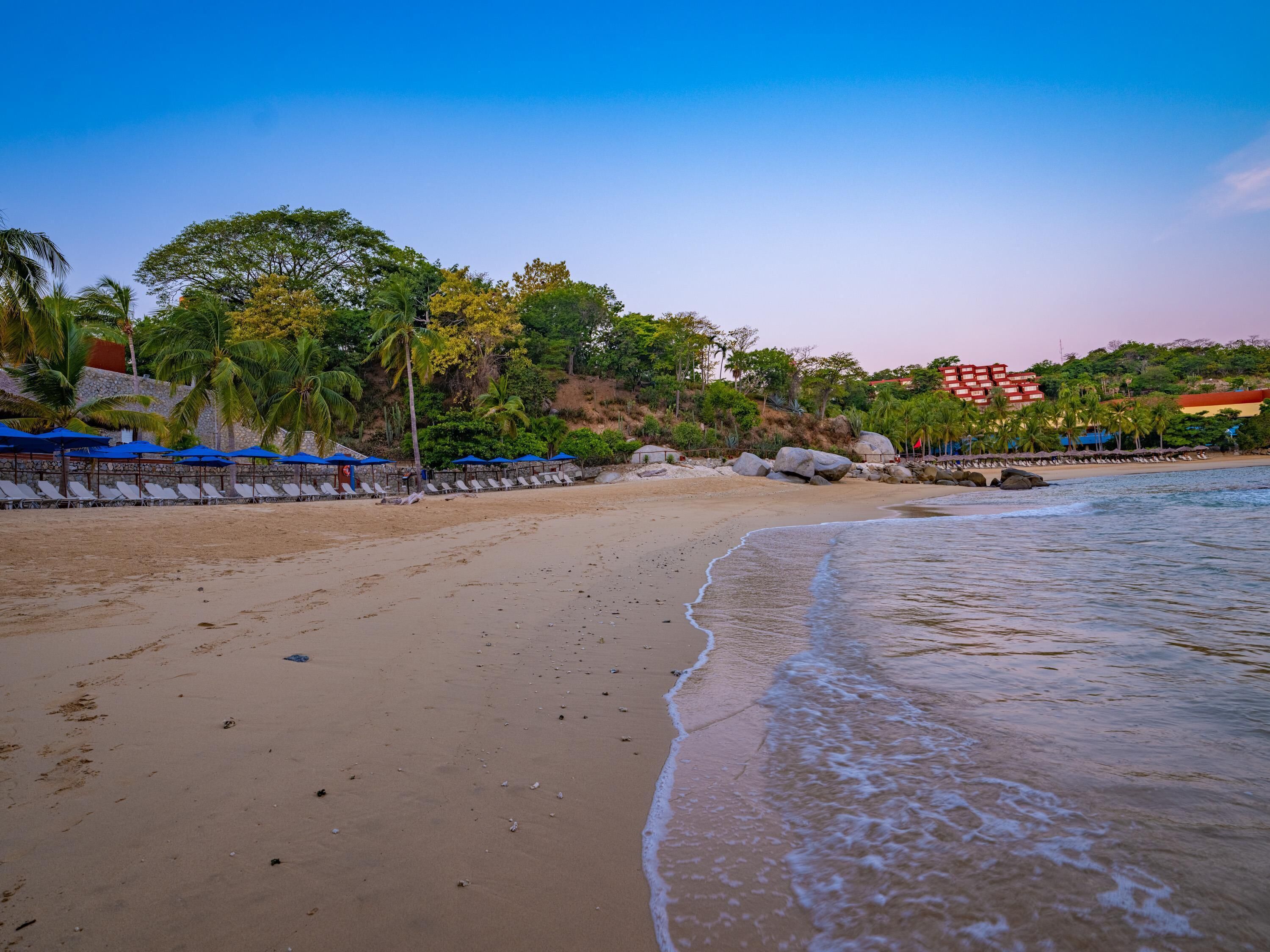 Una playa con filas de tumbonas y sombrillas, un litoral rocoso y un edificio de paredes rojas y amarillas al fondo.