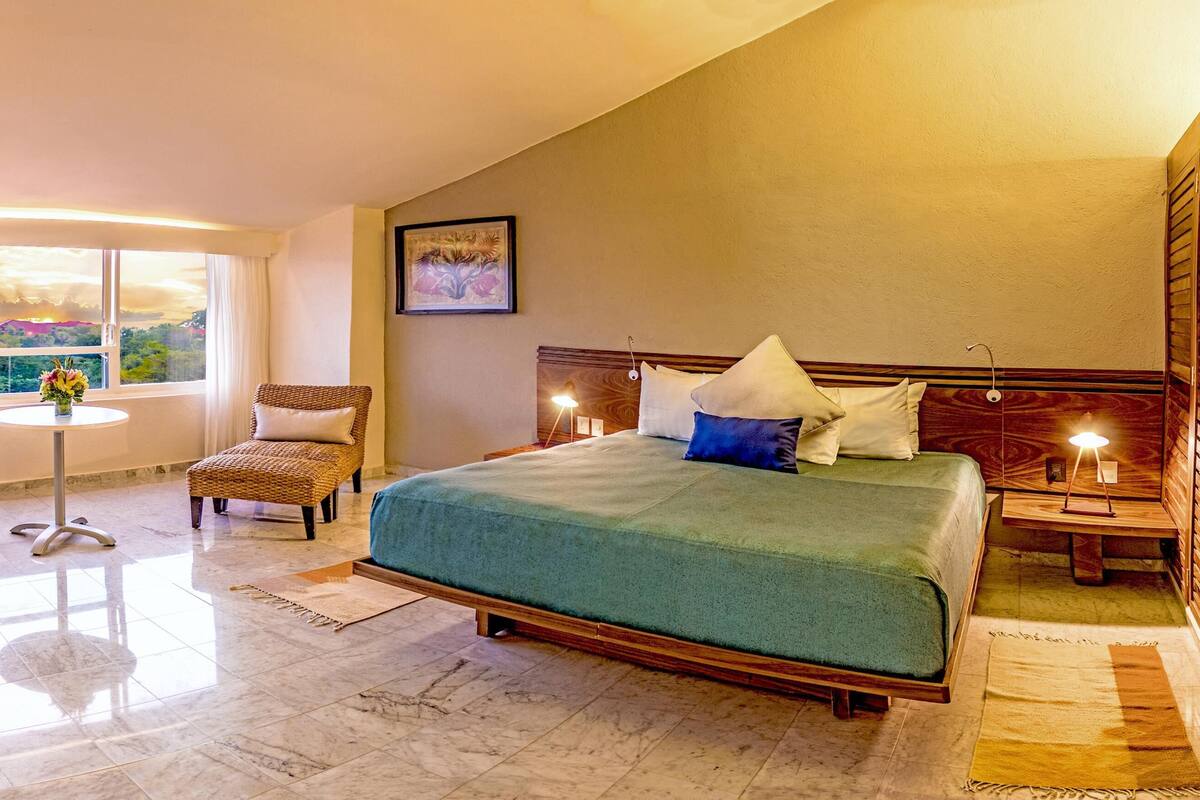 A hotel room with a large bed, a desk, a chair, a small table, and a view of the mountains through the window.