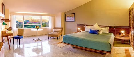 Habitación de hotel con una cama grande, un escritorio, una silla, una mesita y vistas a las montañas desde la ventana.