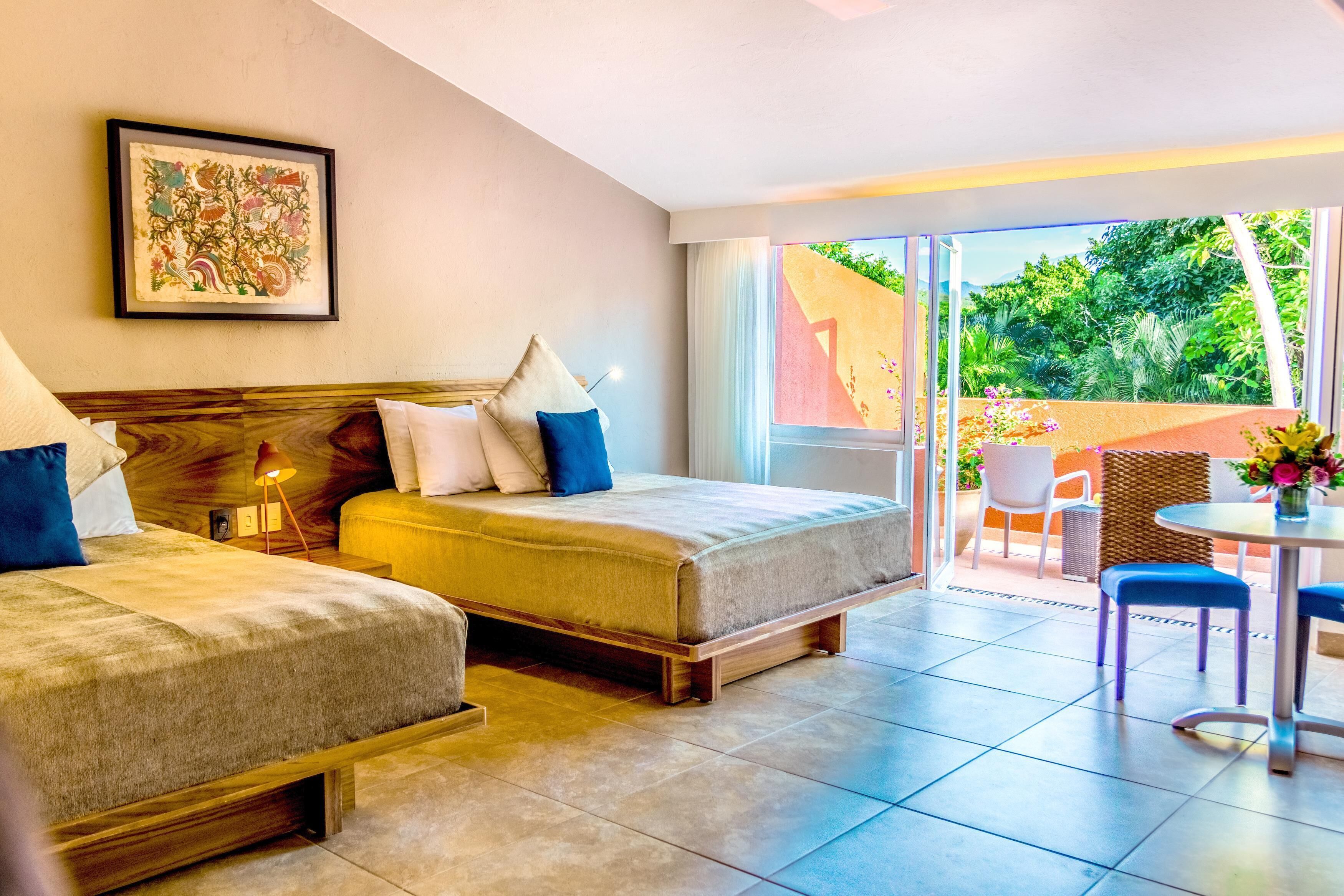 A hotel room with two beds, a balcony with a table and chairs, and a large window offering a view of greenery.
