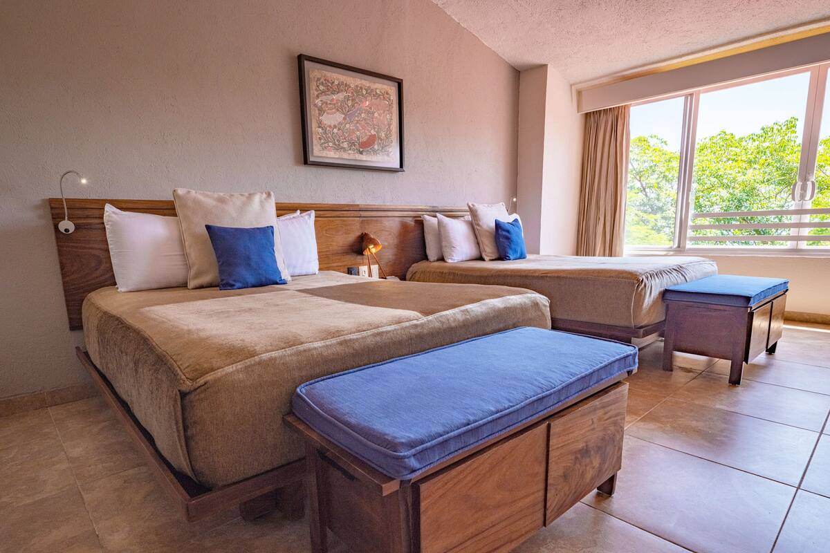 A hotel room with two beds, a wooden headboard, a small table, and a window with a view of trees.