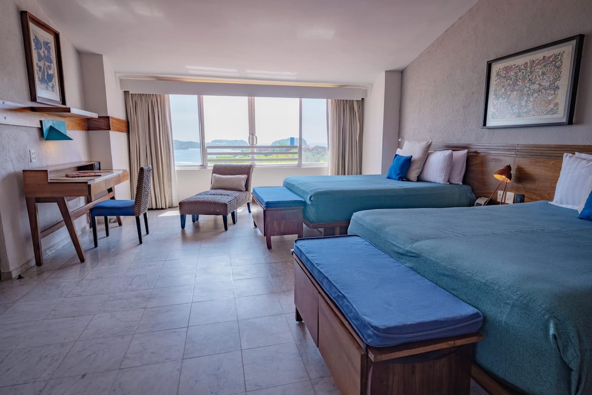 A hotel room with two beds, a desk, a chair, and a view of the sea.