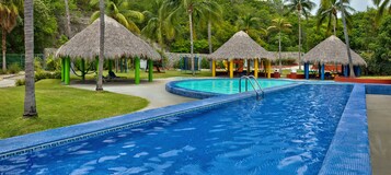 Una piscina con una estructura de techo de paja, rodeada de palmeras y un césped bien cuidado.