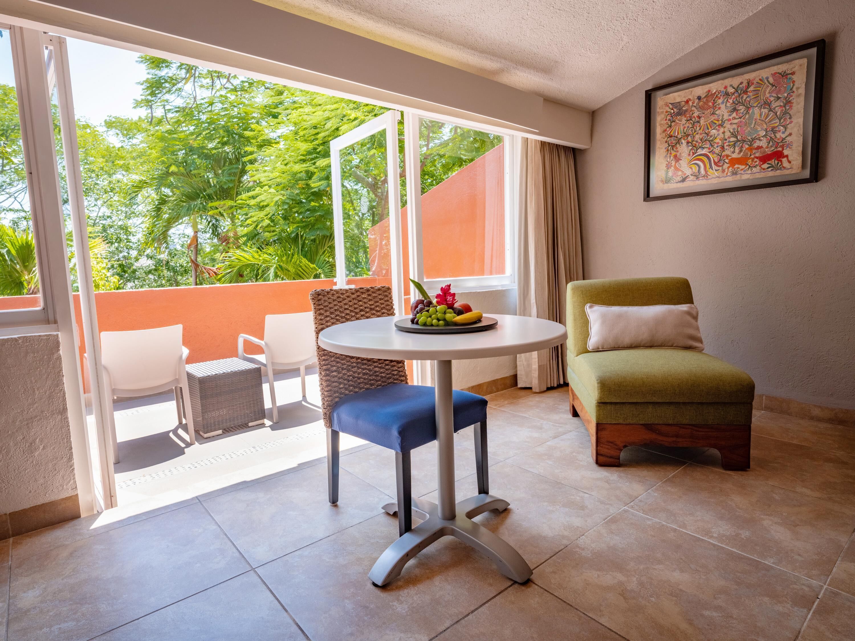 A room with a round table, two chairs, a sofa, and a large window with a view of greenery.