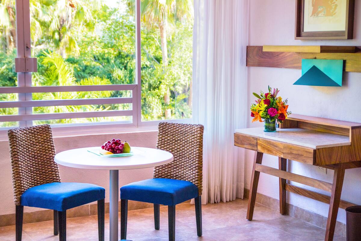 A room with two blue upholstered chairs around a white table, a wooden desk, and a window with a view of greenery.