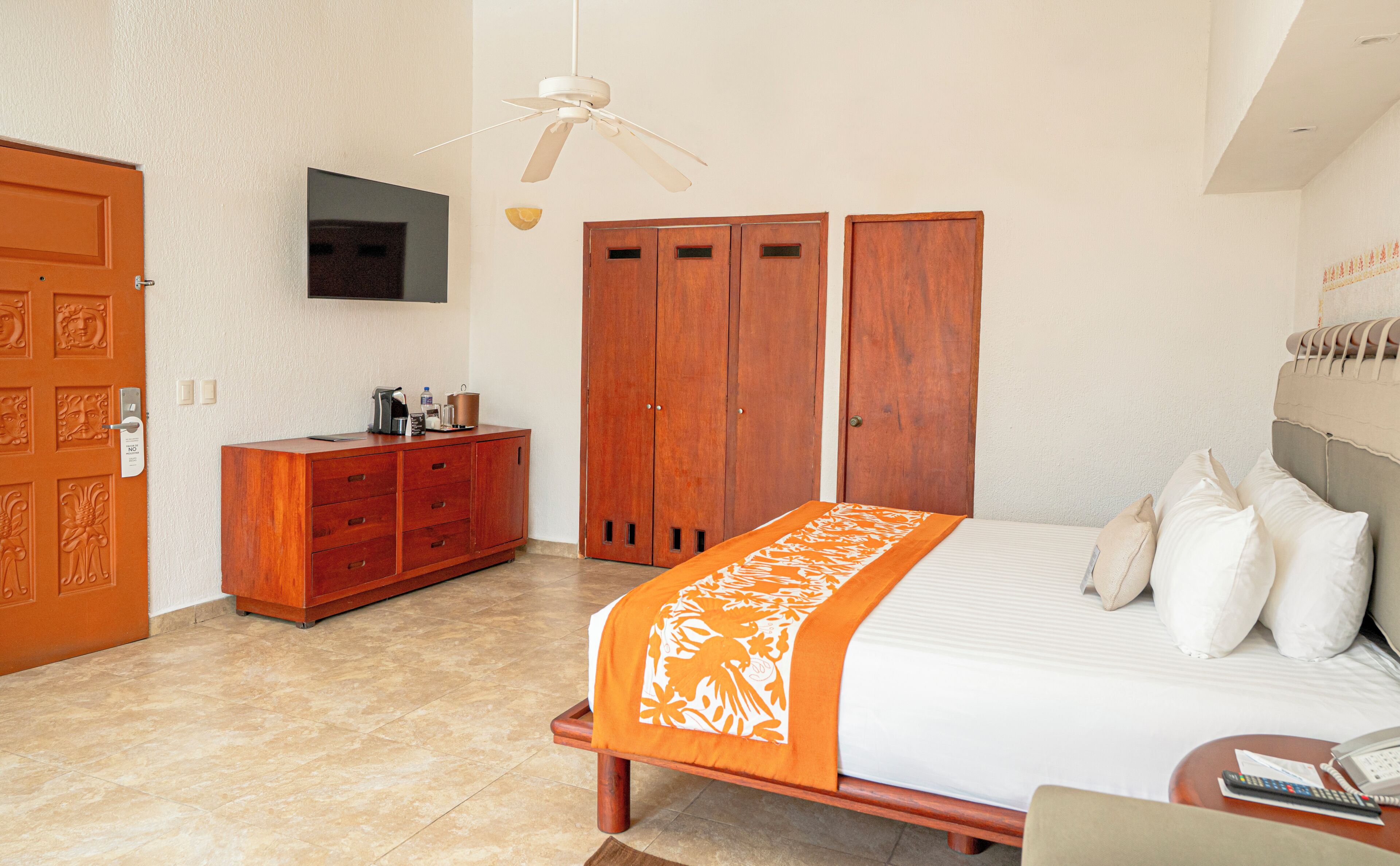A bedroom with a bed, a nightstand, a wooden wardrobe, a ceiling fan, and a television on the wall.
