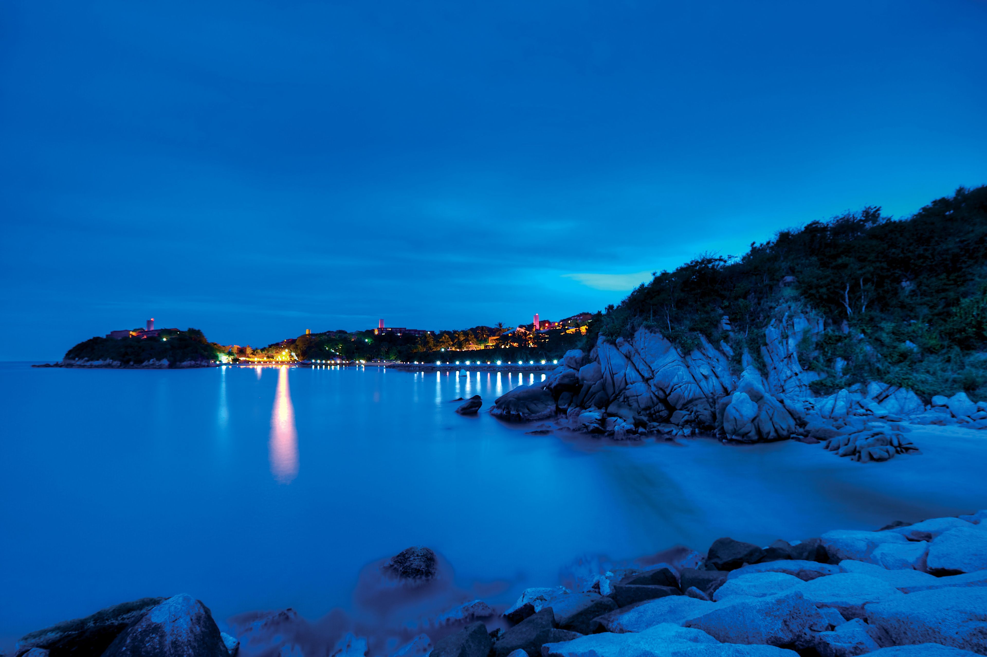 A coastal scene with a calm sea, rocky shoreline, and a distant illuminated town.