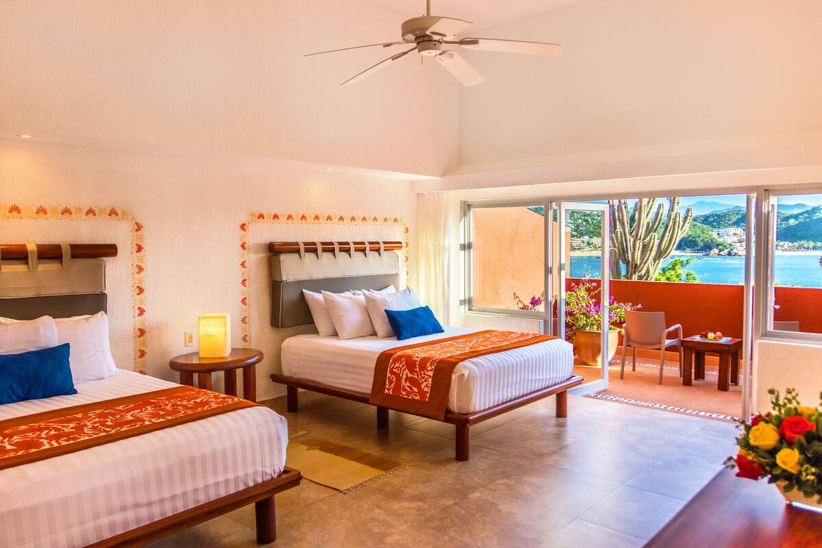 A hotel room with two beds, a ceiling fan, a balcony with a view of the sea, and a small table with a vase of flowers.