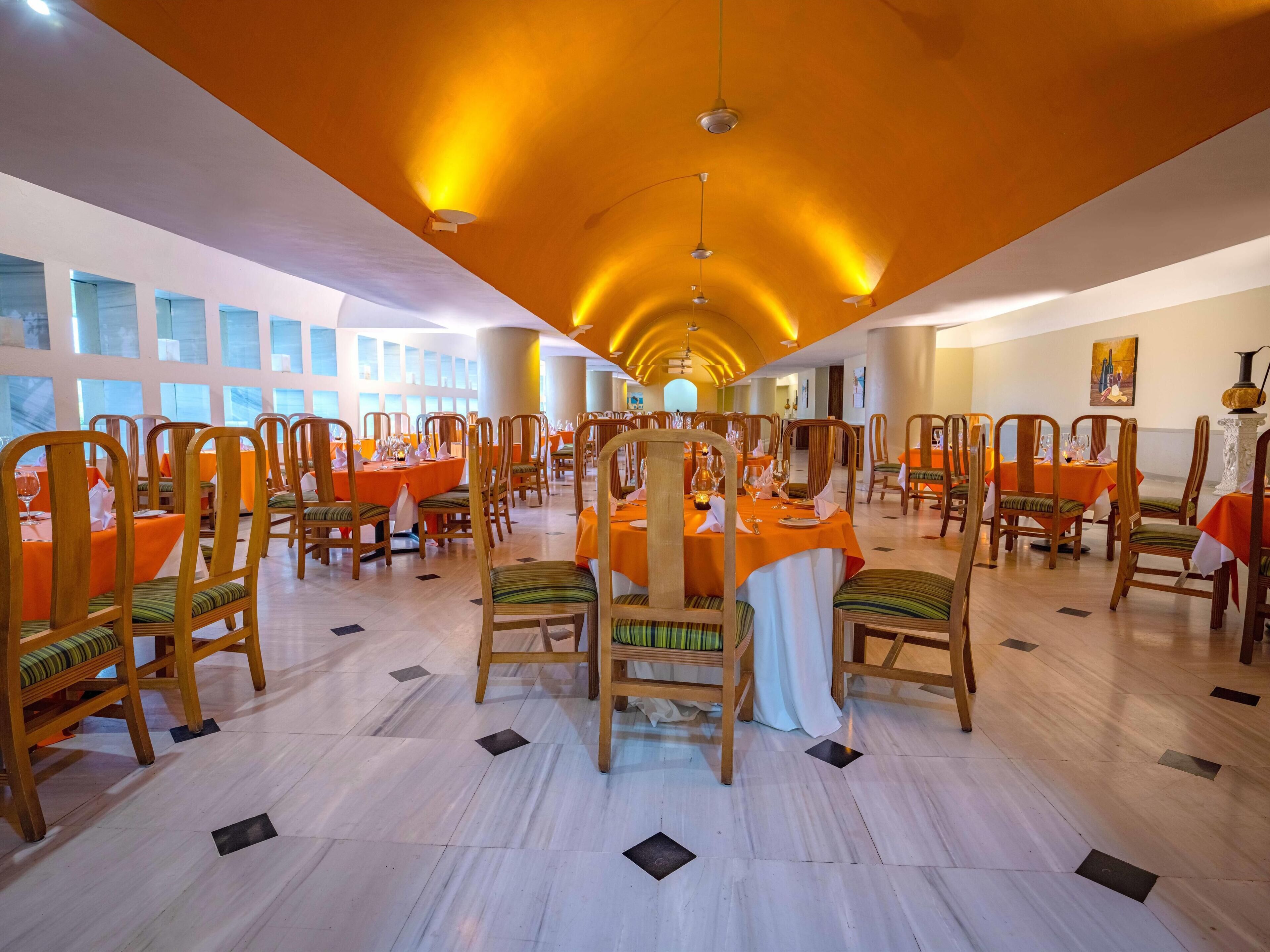 A spacious dining hall with multiple tables and chairs, large windows, and a high ceiling with recessed lighting.