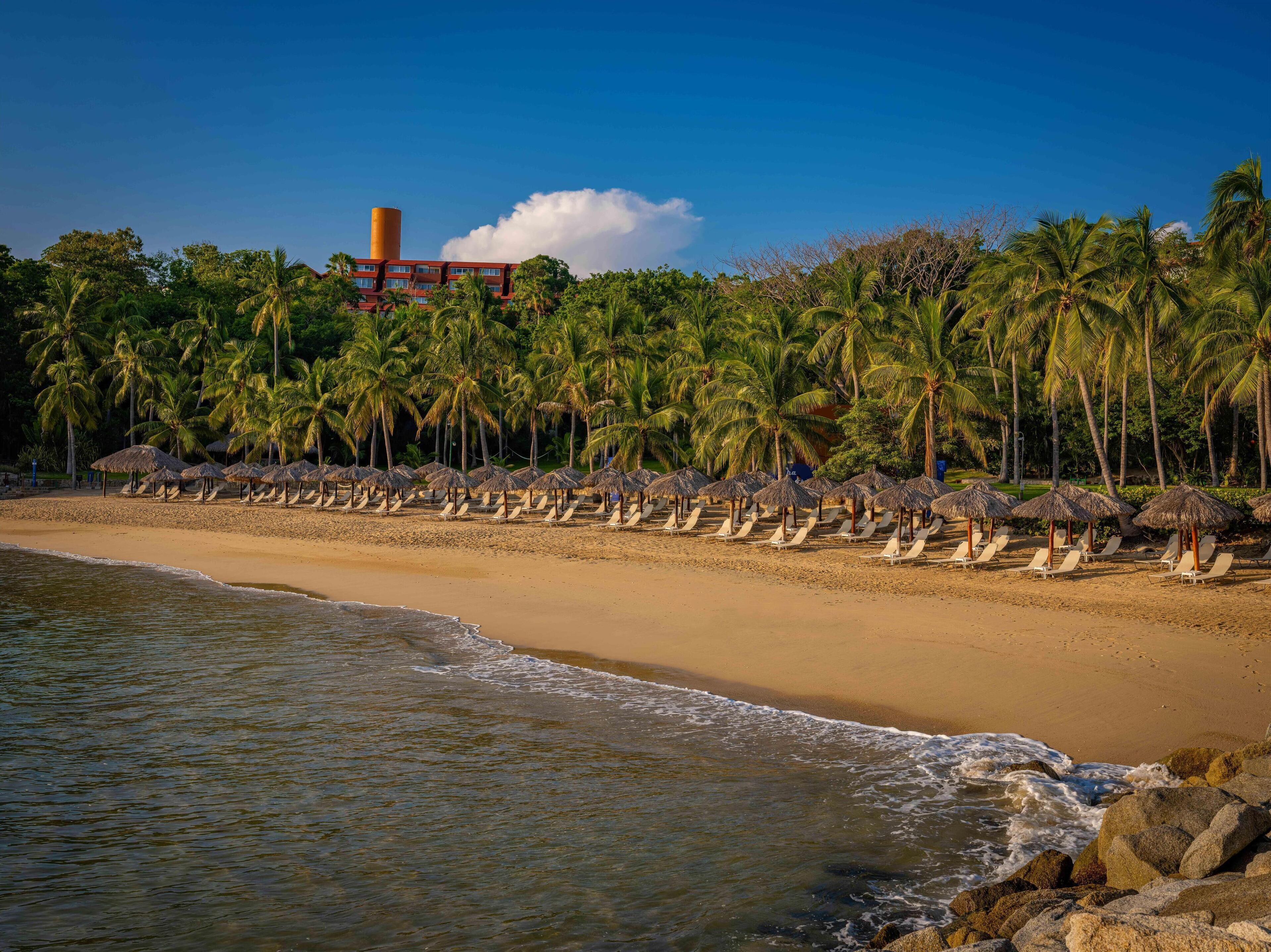A beach with rows of sun loungers and thatched umbrellas, palm trees, and a building with a smokestack in the background.