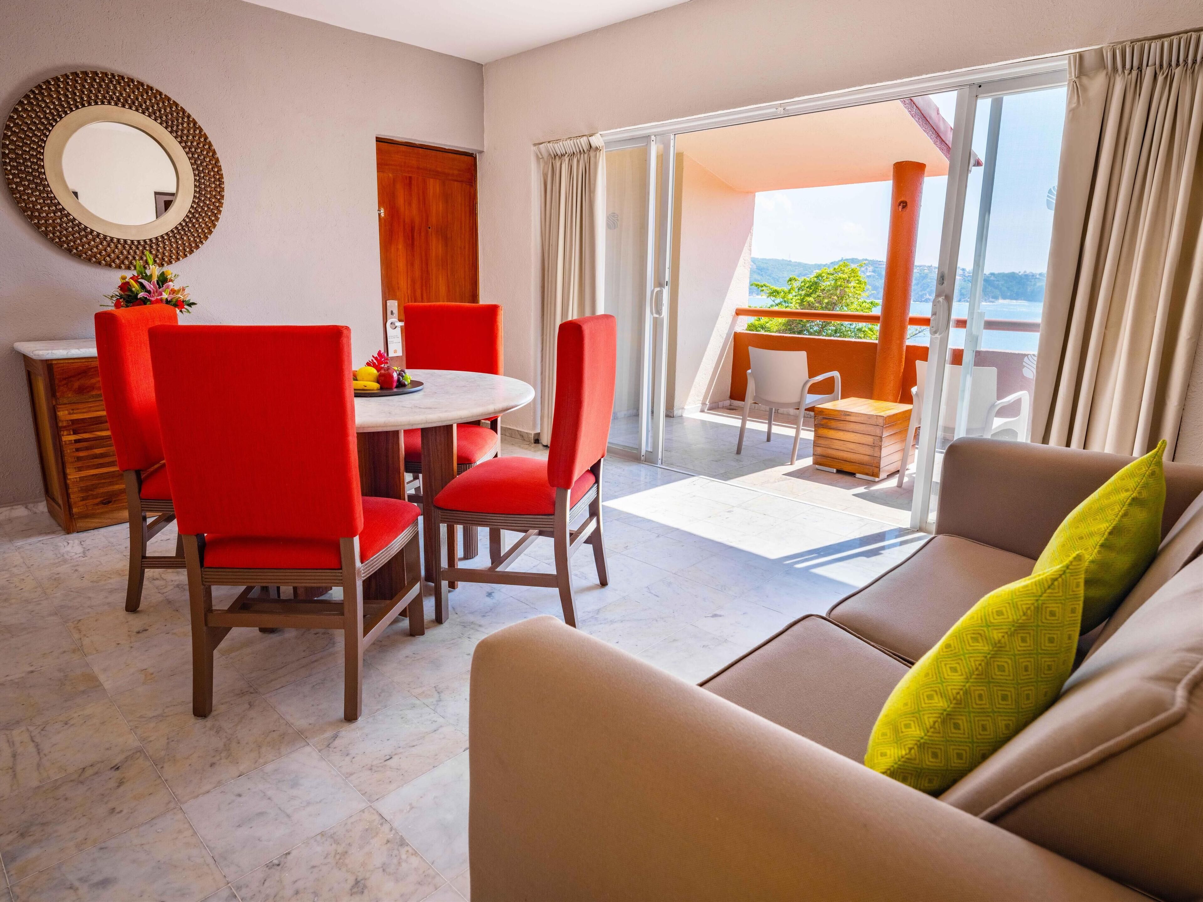 A modern living room with a round table, red chairs, a beige sofa, and a balcony with a view.