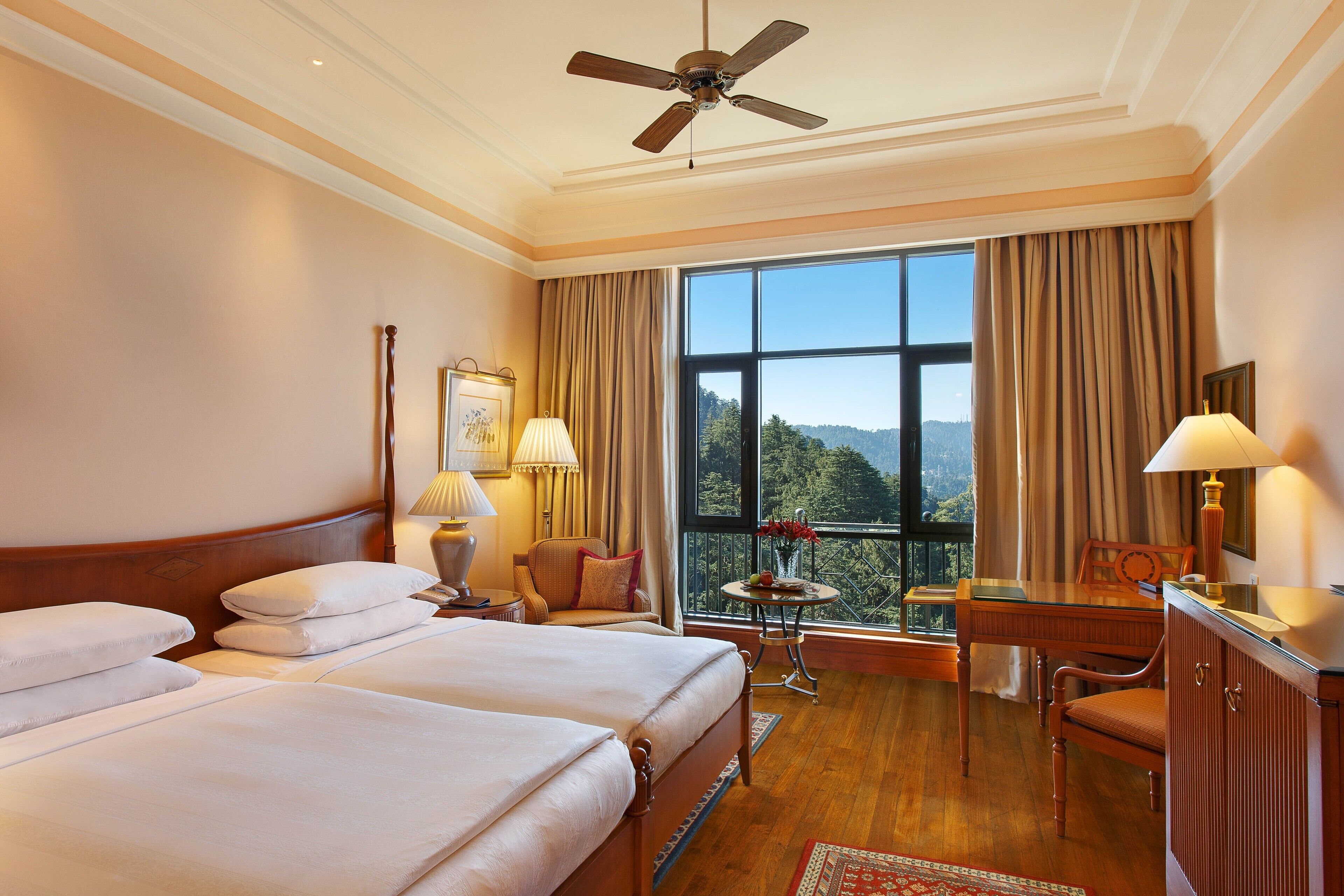 Habitación de hotel con dos camas, un escritorio, una silla, una mesita con lámpara y vistas al exterior.