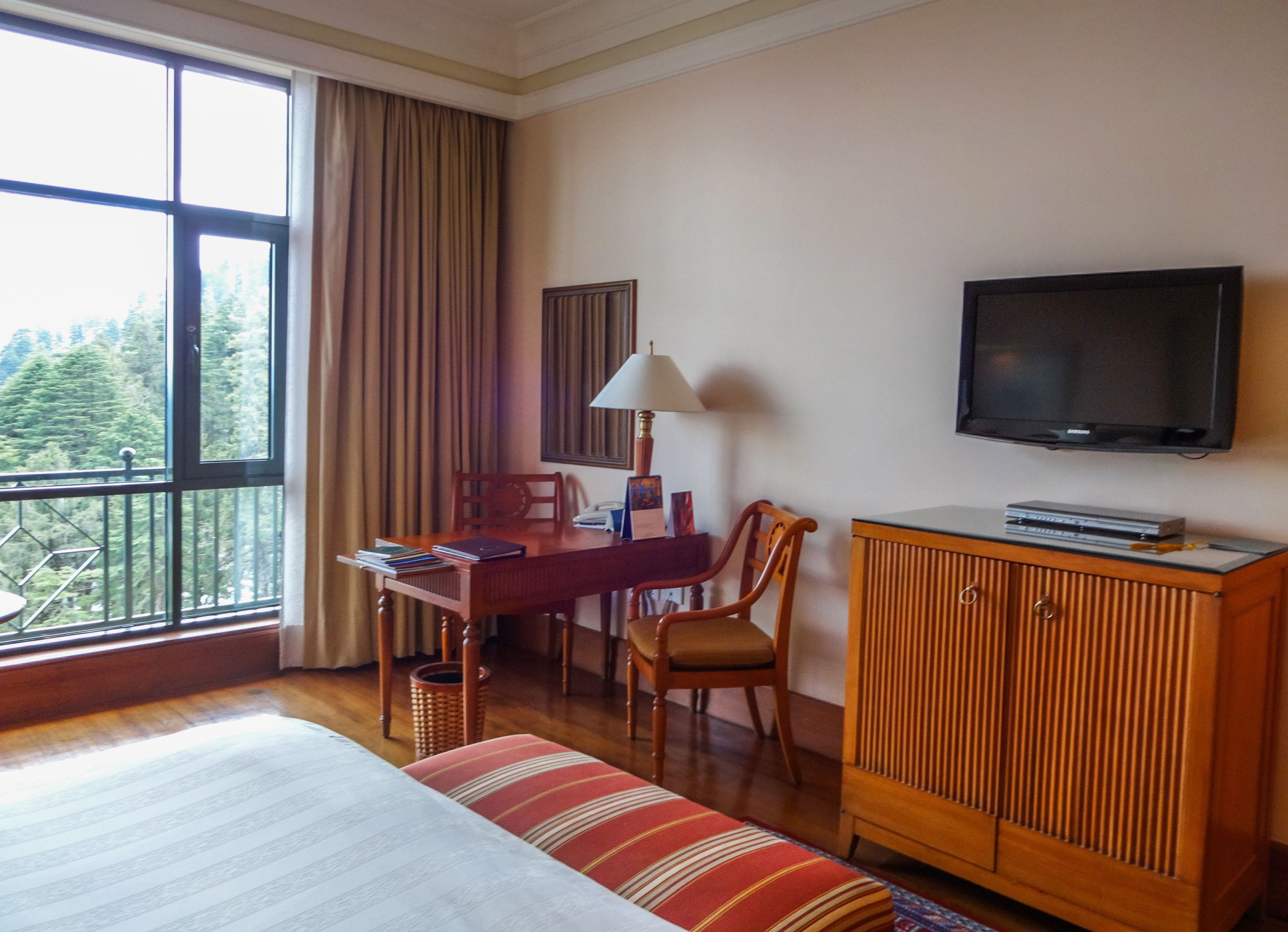 Habitación de hotel con cama, escritorio con lámpara, televisor y vista a un bosque a través de un ventanal grande.